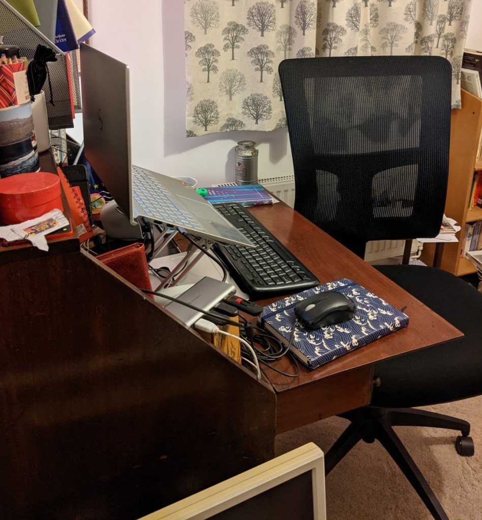 Bureau style desk with a laptop on a raiser, keyboard, mouse, etc, and an adjustable chair