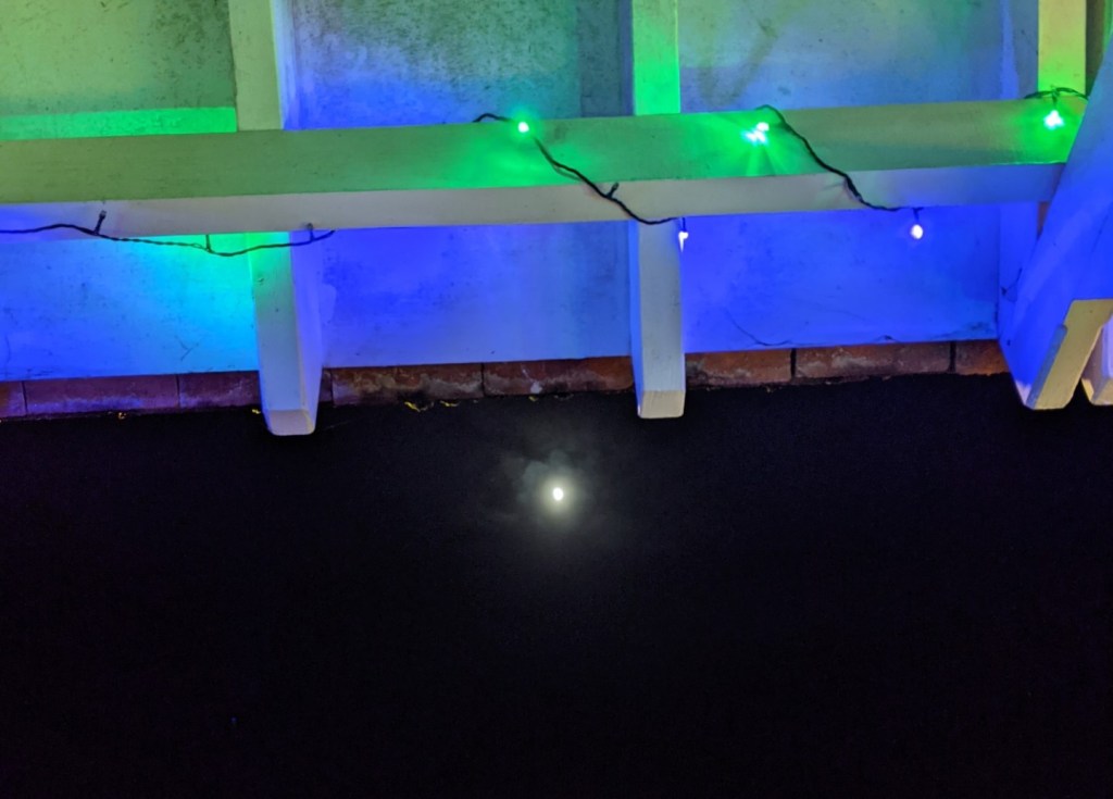 waxing gibbous moon seen just below the edge of a wooden porch, which has blue and green fairy lights twined around the beam
