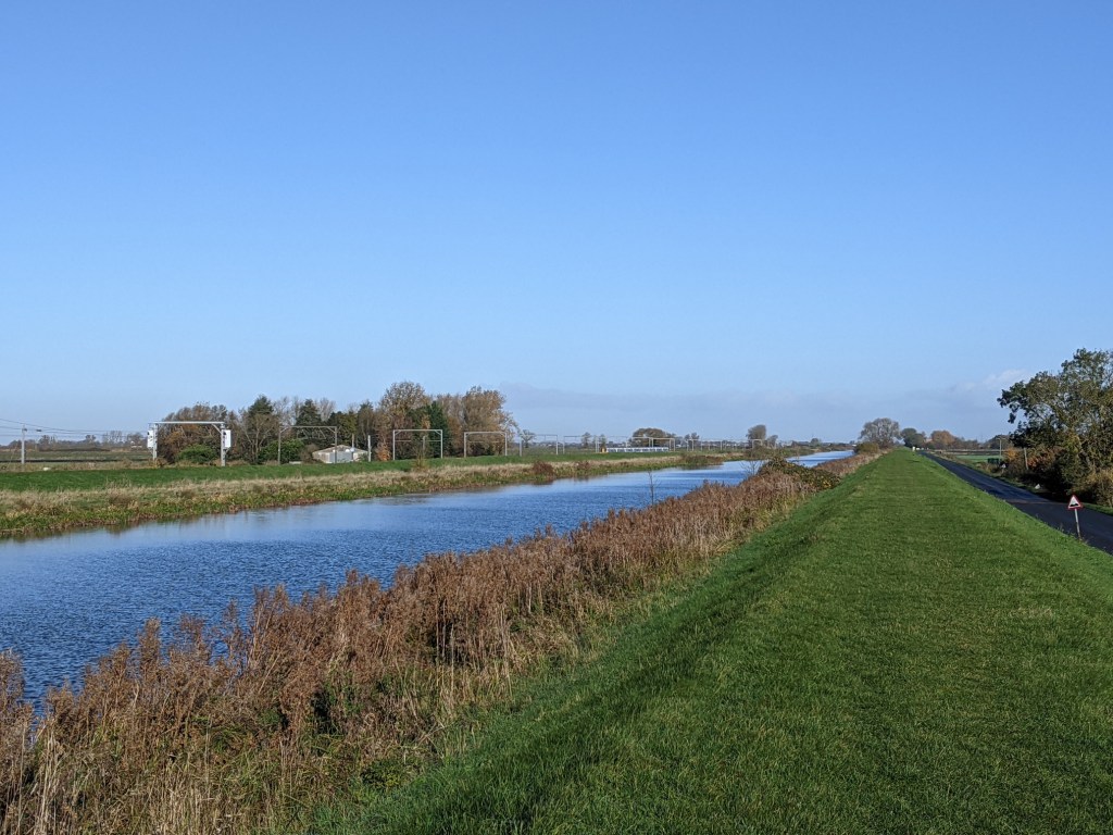 Blue sky reflected in a straight, calm, river, with reeds and a grassy artificial dike on the right bank and overhead railway power lines on the left bank
