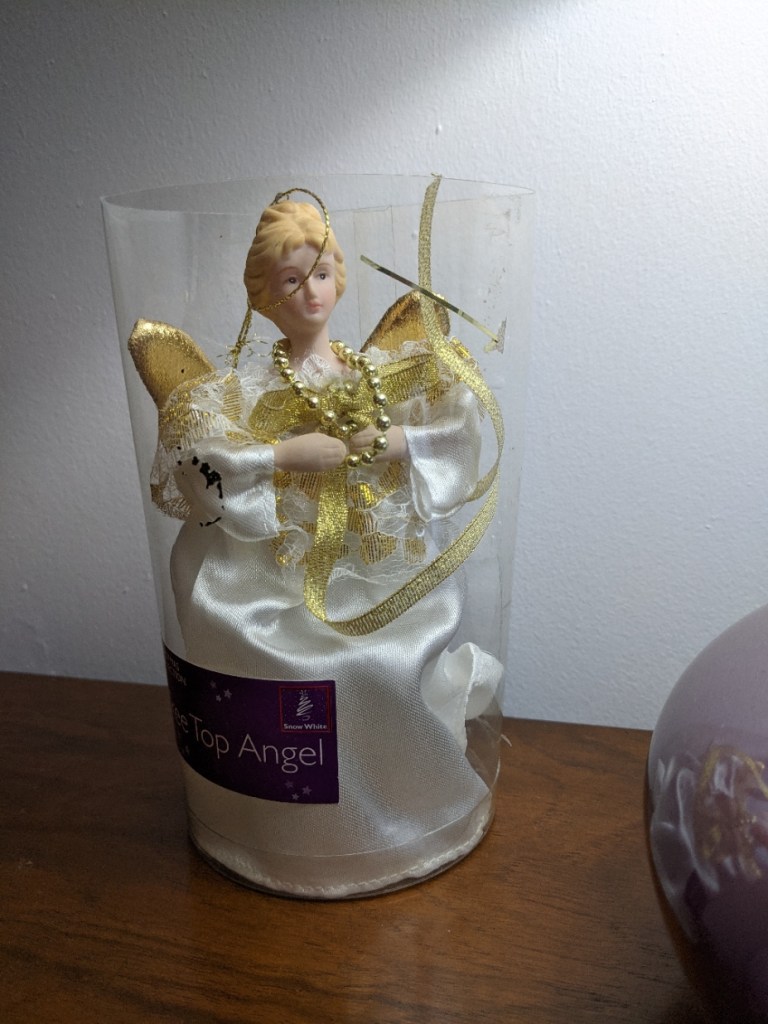 A Christmas decoration representing an angel, in a plastic tube. She has ceramic head an hands, gold fabric wings, and a white satin robe.