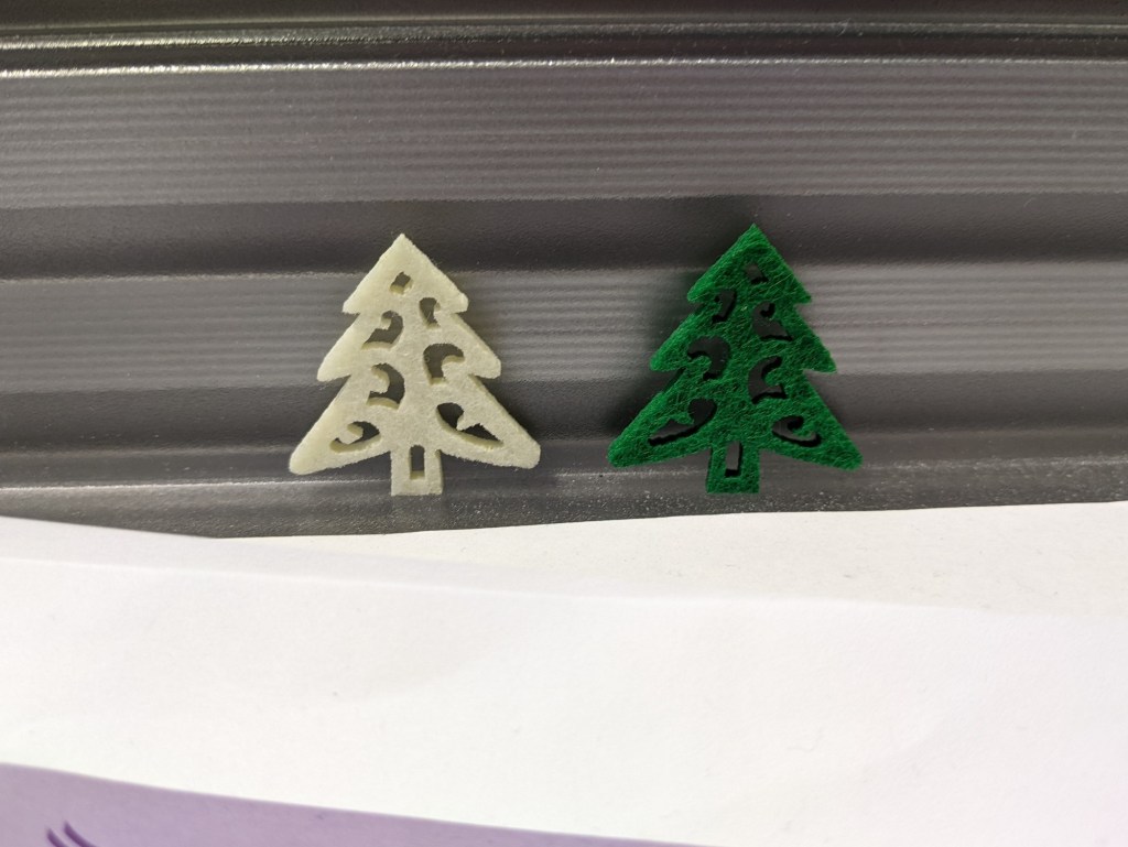Two tiny felt cut-outs, one cream and one dark green, in the shape of Christmas trees, standing in a transparent plastic tray