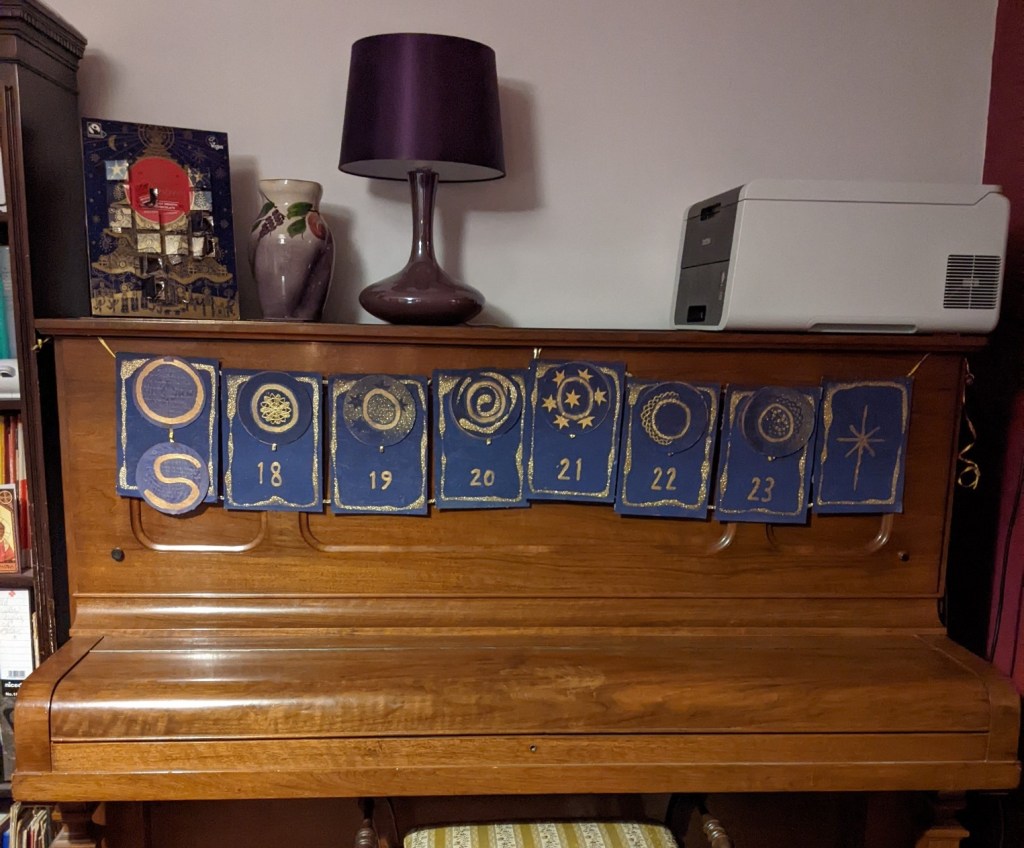 Eight purple cards strung across the front of an upright piano. All but one of them has a circle of card with the letter O on it. The right hand most card only has a star painted on it. On the left hand most card the circle has been opened to reveal a second one, whcih has a letter S.