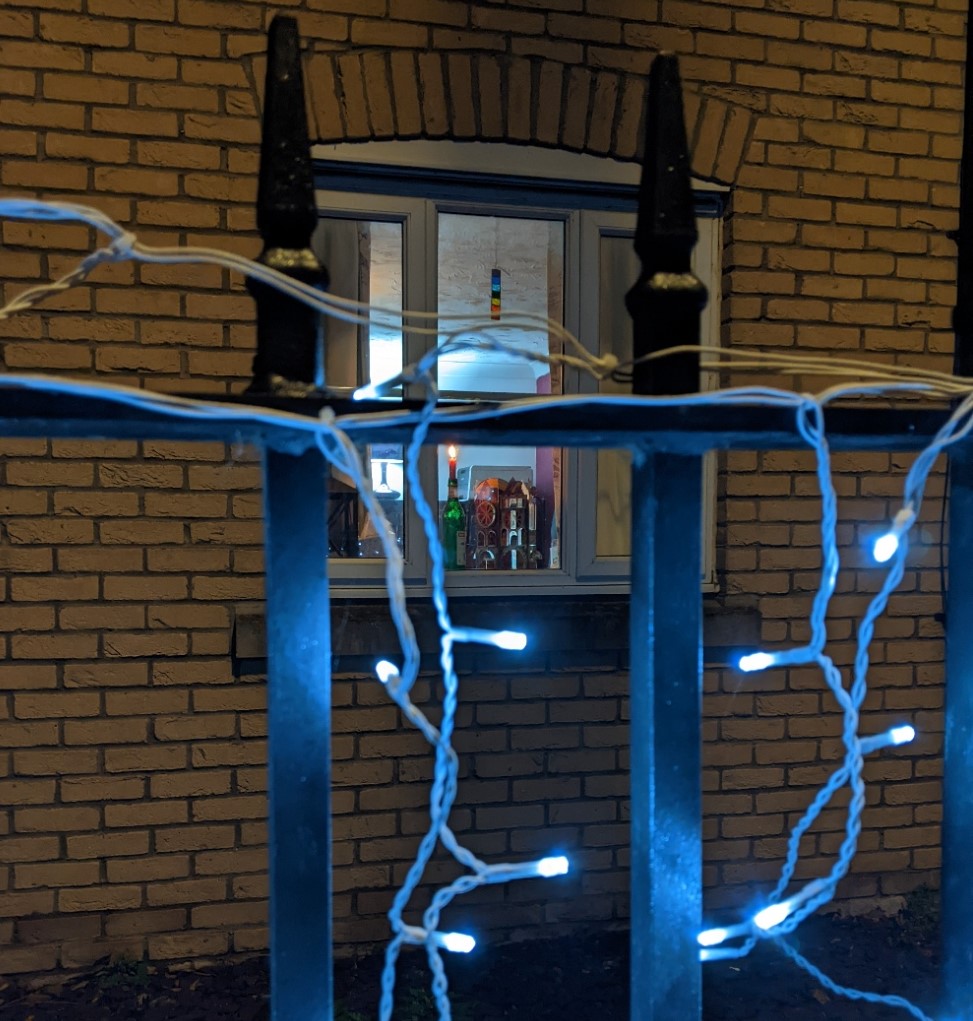 Metal railings with blue-white fairy lights twined around them and cascading down them. In the background a candle is burning in a window