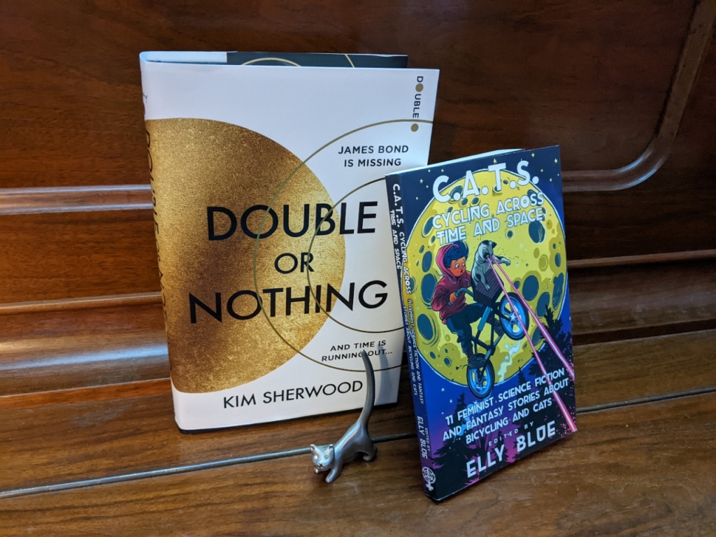 Hardback copy of 'Double or Nothing' by Kim Sherwood, paperback copy of 'CATS: Cycling Across Time and Space' edited by Elly Blue, small metal cat with crystal eyes
