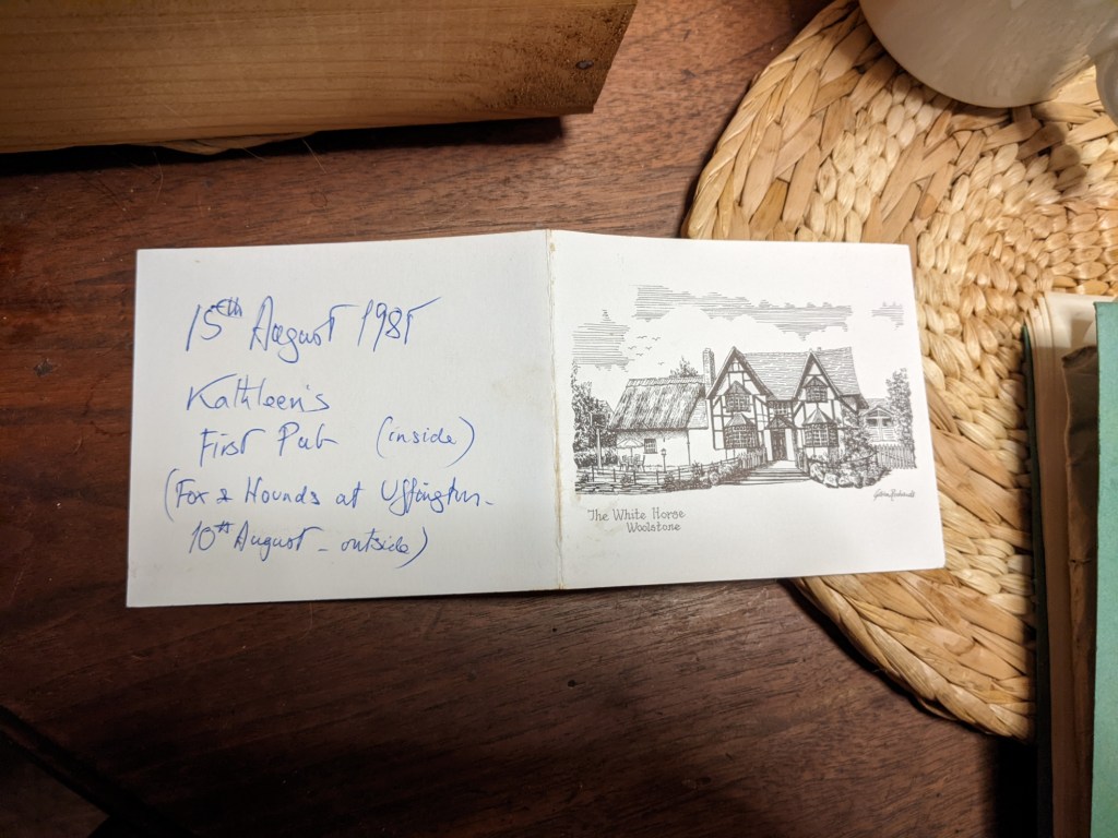 A business card from The White Horse at Woolstone, with handwritten text on the back:
15th August 1985
Kathleen's First Pub (inside)
(Fox & Hounds at Uffington - 10th August - outside)