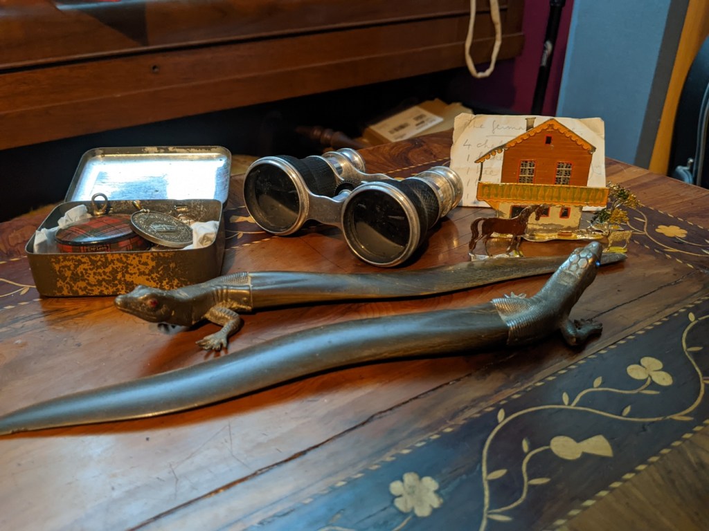 Collection of objects on a wooden table: two letter openers shaped like lizards; a pair of opera glasses; tin containing a medal with the image of the Royal Albert Hall; metal chalet, horse, tree and dog