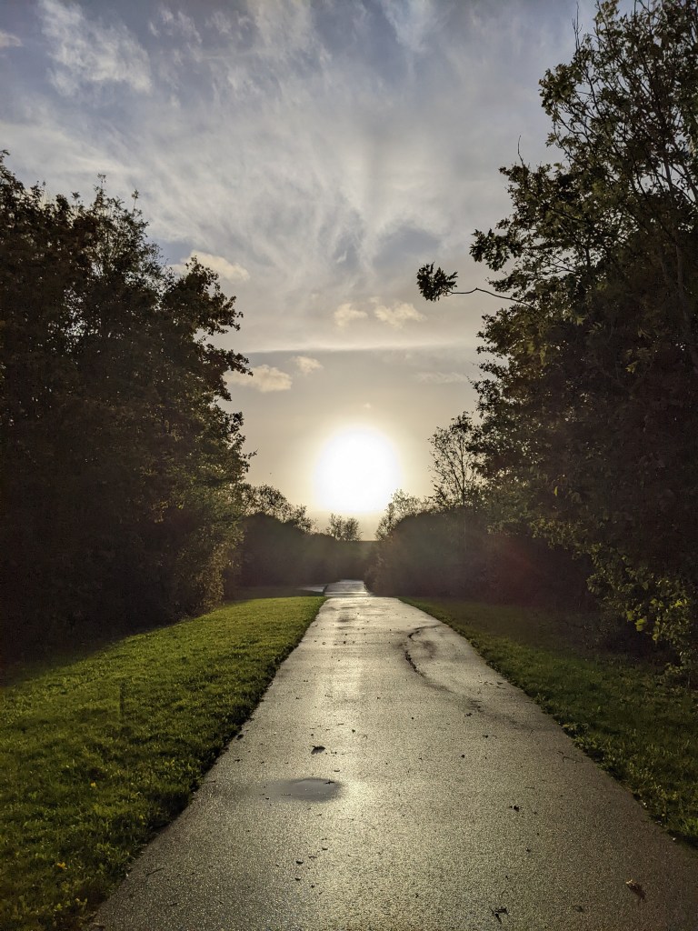 Looking down a wet tarmac path into a bright pale yellow sunset 