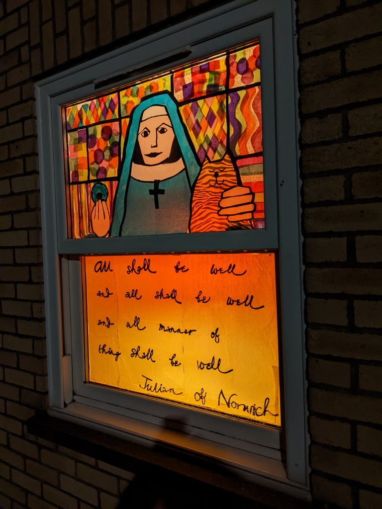 House window decorated with tissue paper to make a picture of a medieval religious woman holding a small circular object in one hand and embracing a striped cat with the other. Text underneath reads "All shall be well and all shall be well and all manner of thing shall be well. Julian of Norwich"