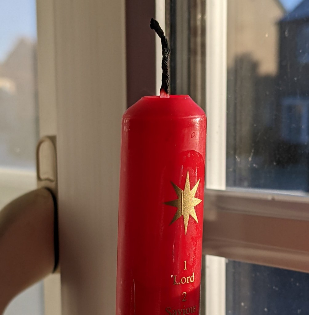 A red candle which has been lit and extinguished, with gilt decoration. Visible in this photo are an eight pointed star, a figure 1 and the word Lord. It is daylight.