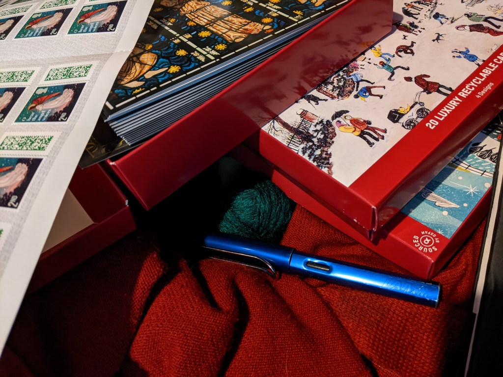 Boxes of cards, one featuring stained glass angels and one with a winter street scene, red knit fabric, stamps, and a blue fountain pen