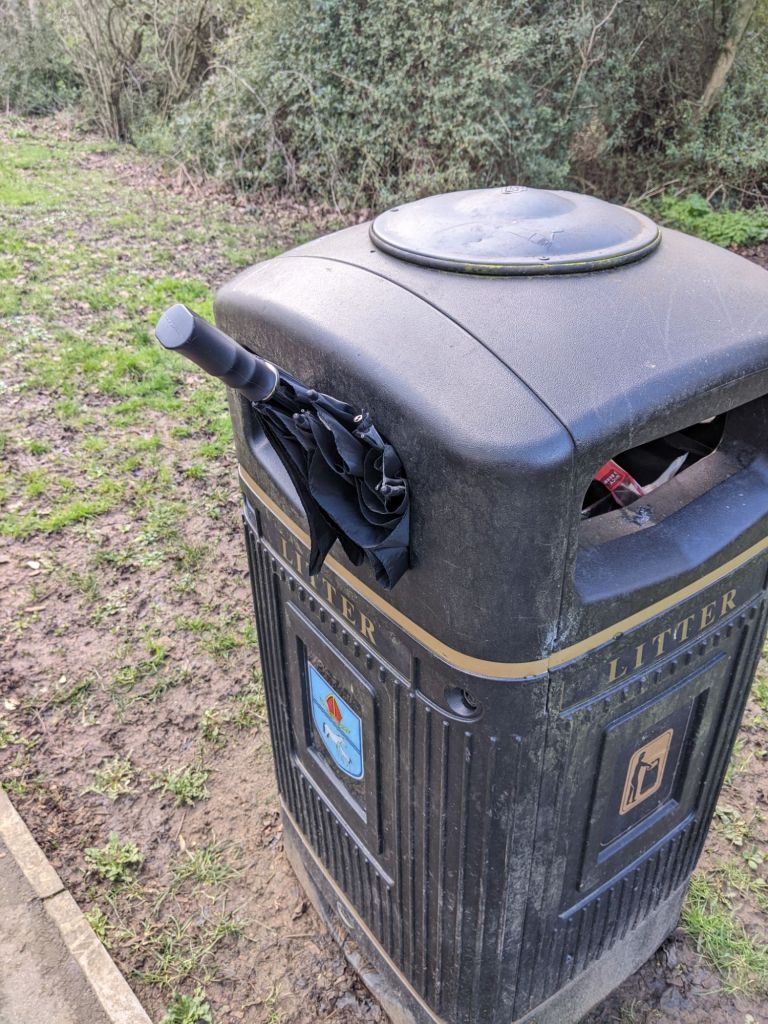 A litter bin with the handle end of a black umbrella poking out of it