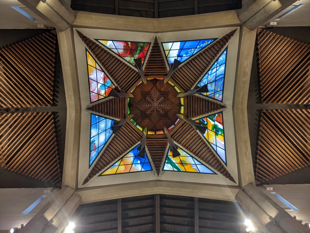 Looking upwards into a church ceiling where wood and concrete form an eight-pointed star, with the gaps filled with blue, yellow, green and red stained glass