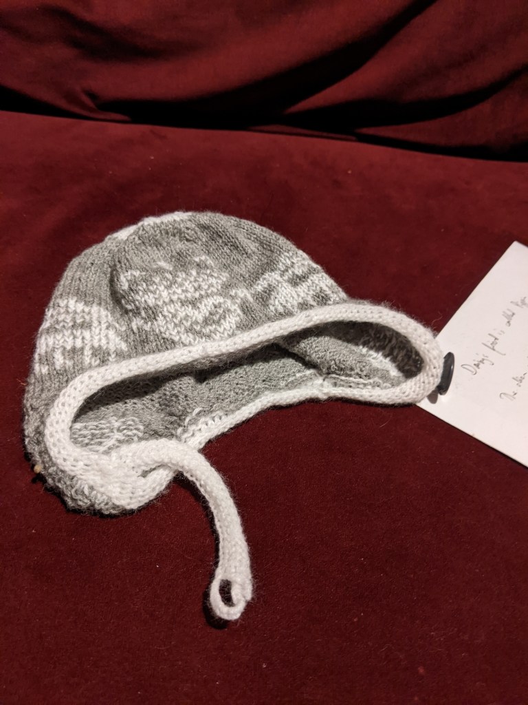 A baby's knitted hat in grey yarn with a white trim and a pattern of deer and a crowned heart in white 