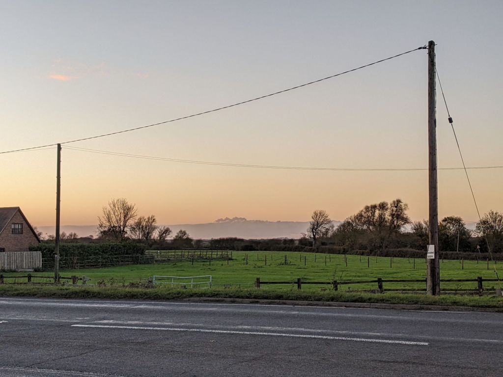View across flat grassy fields. In the background a low bank of cloud bears a remarkable resemblance to distant mountains