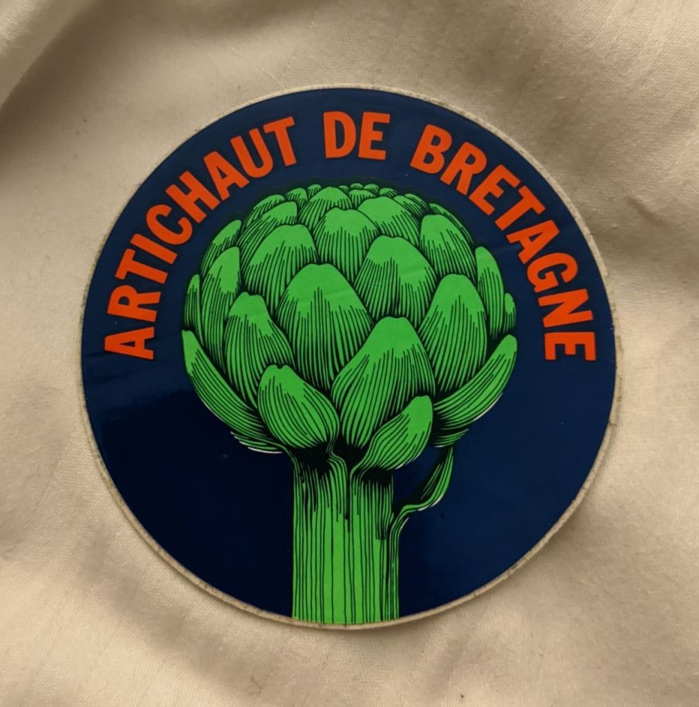 Vinyl sticker, still on its backing paper, with a bright green globe artichoke on a dark blue background with text 'Artichaut de Bretagne ' in red