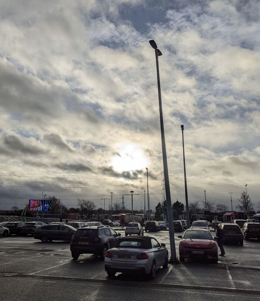 Across a car park, the low sun shines through grey clouds, though there's a bit of blue sky higher up