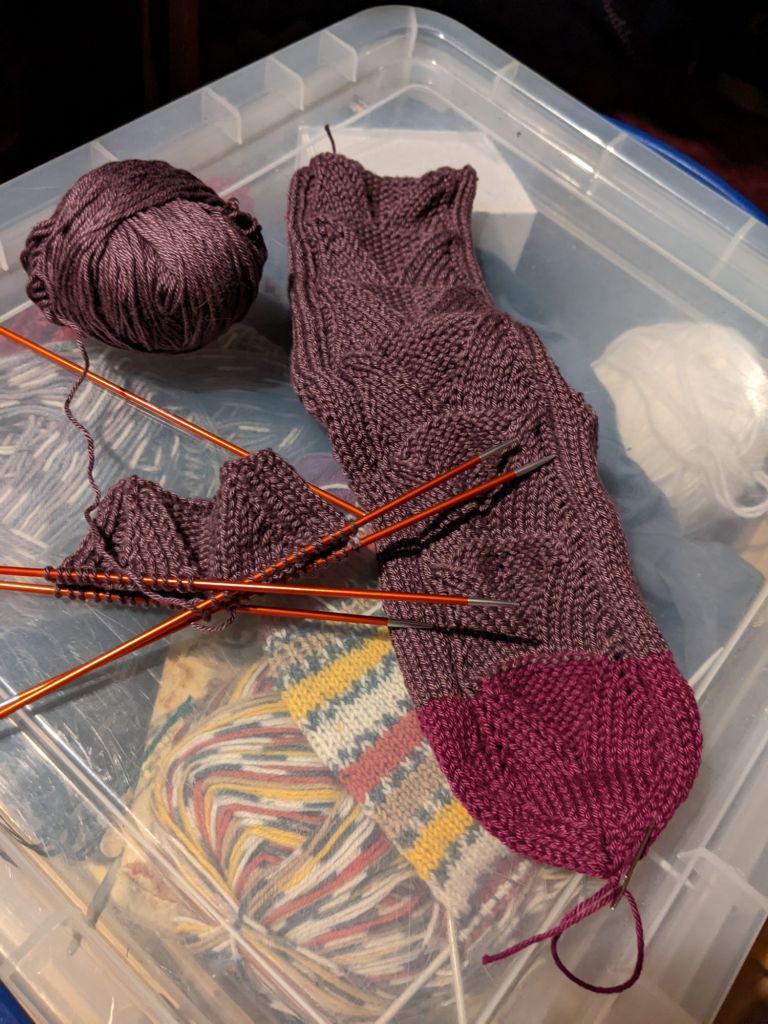 Knitted sock in two shades of purple. The beginning of the second sock is still on the knitting needles.
