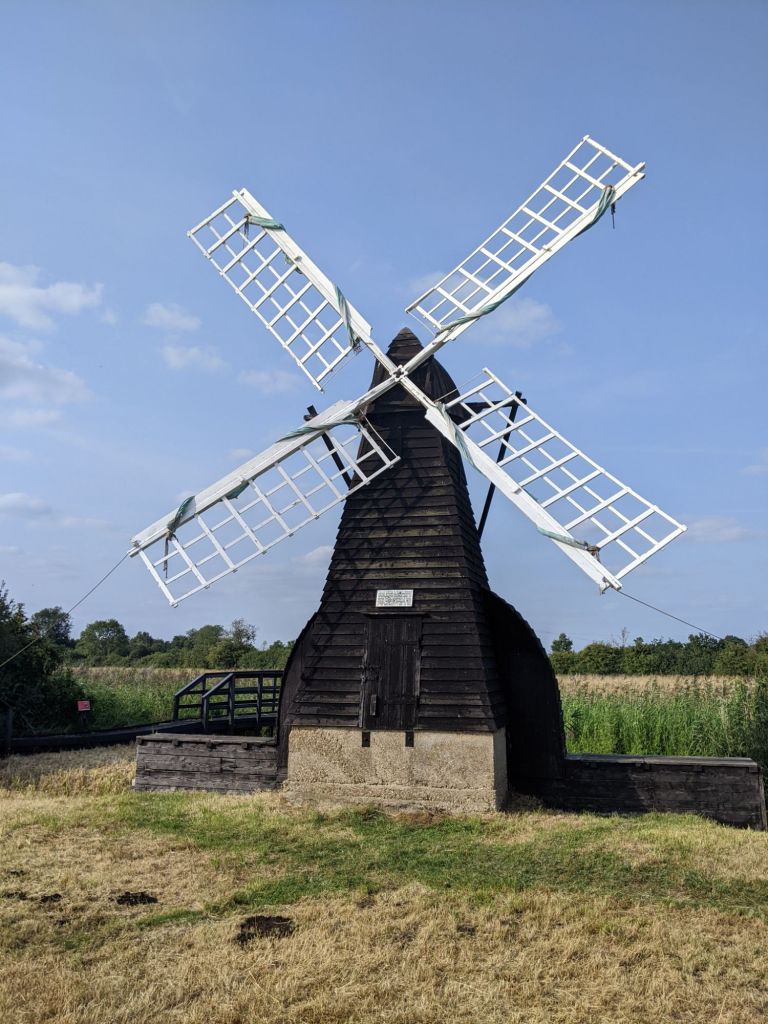 Windpump, main structure of dark wood planks, white sails, against a blue sky 