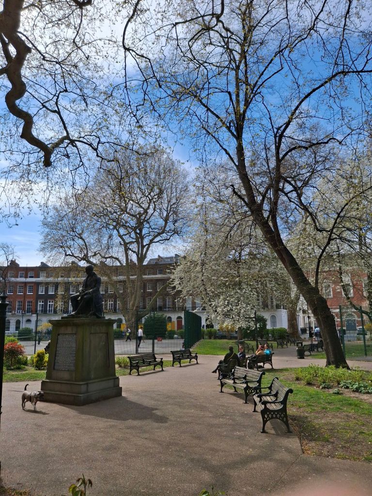 A London public garden on a sunny spring day, with trees and tennis courts and a statue of a seated luminary