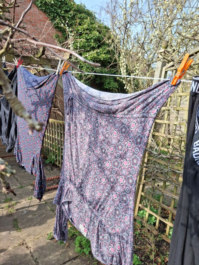 Clothes with floral patterns in purple, black, deep blue colour schemes hang on a washing line. In the background a fruit tree is just beginning to bloss6