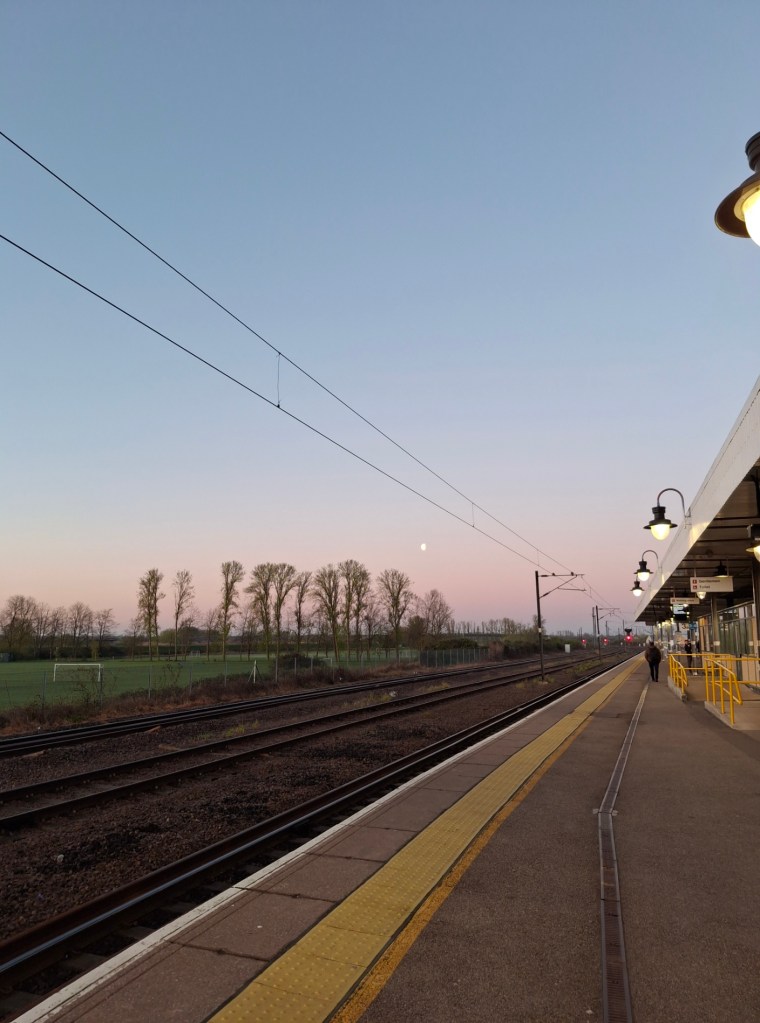 A railway station early in the morning, with a clear blue sky shading to pink at the horizon, and a half-moon low in the sky above bare trees