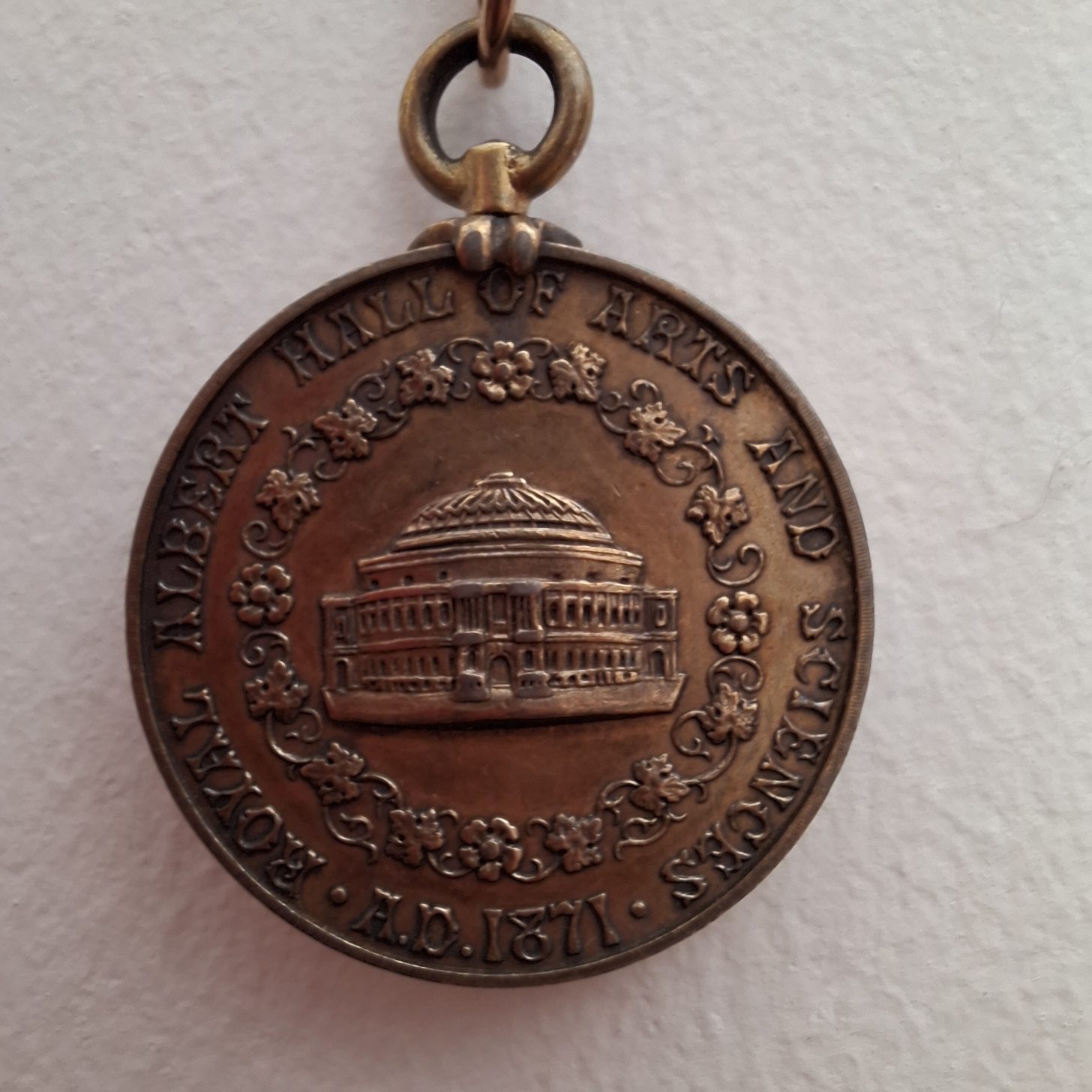 Bronze-coloured medal depicting the Royal Albert Hall surrounded with a garland of roses and vine leaves and text "Royal Albert Hall of Arts and Sciences A. D. 1871"