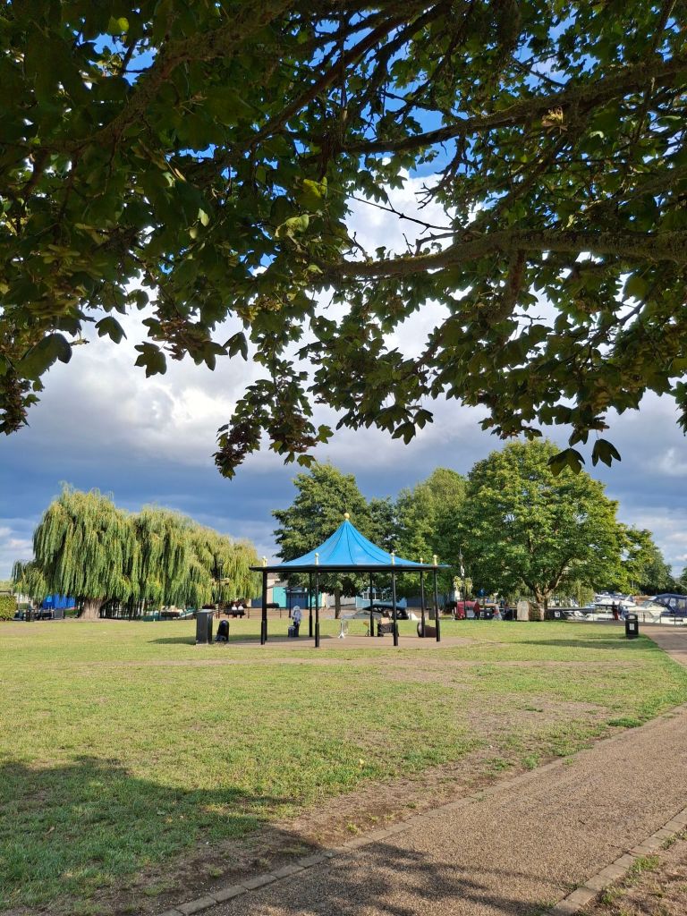 A circular bandstand with a blue roof stands in an almost deserted park. Under the bandstand there is one lone tuba standing upside down on its bell and one lone human