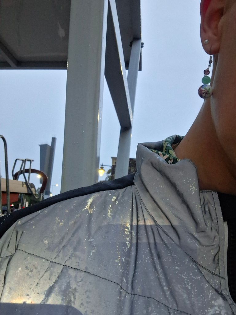 The shoulder and part of the face of someone wearing a silvery reflective cycling jacket, with a light sprinkling of raindrops. Behind them can be seen a double decker cycle storage rack and grey sky