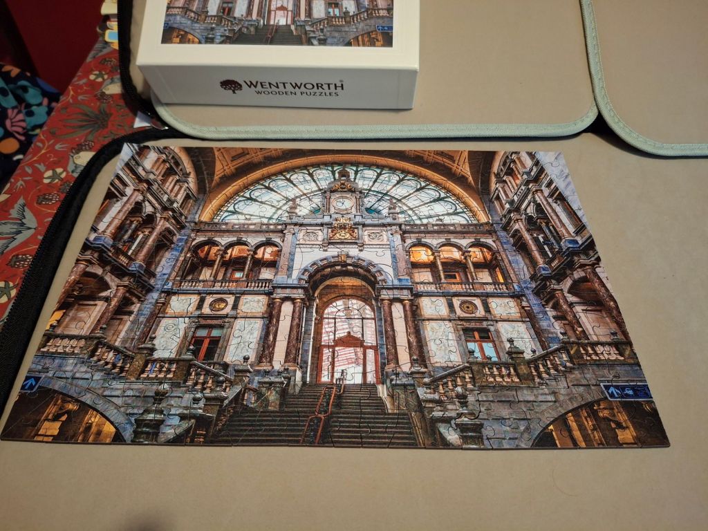 A completed jigsaw puzzle, next to its box. The image is a railway station hall with a lot of grey stone columns, a huge semi-circular window at the top, and some gilt decorations. 