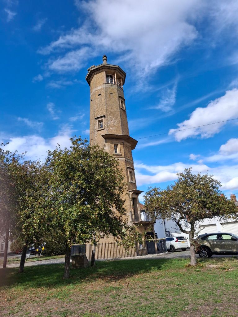 Tall lighthouse in grey-brown brick. It's a little way inland so there are trees, grass and cars around the base.