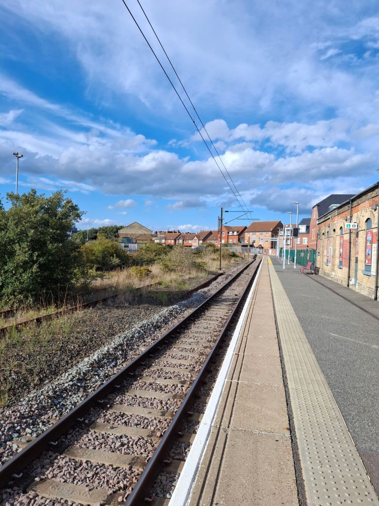 Single track railway line running alongside a single storey station building and coming to a stop at a wooden fence 