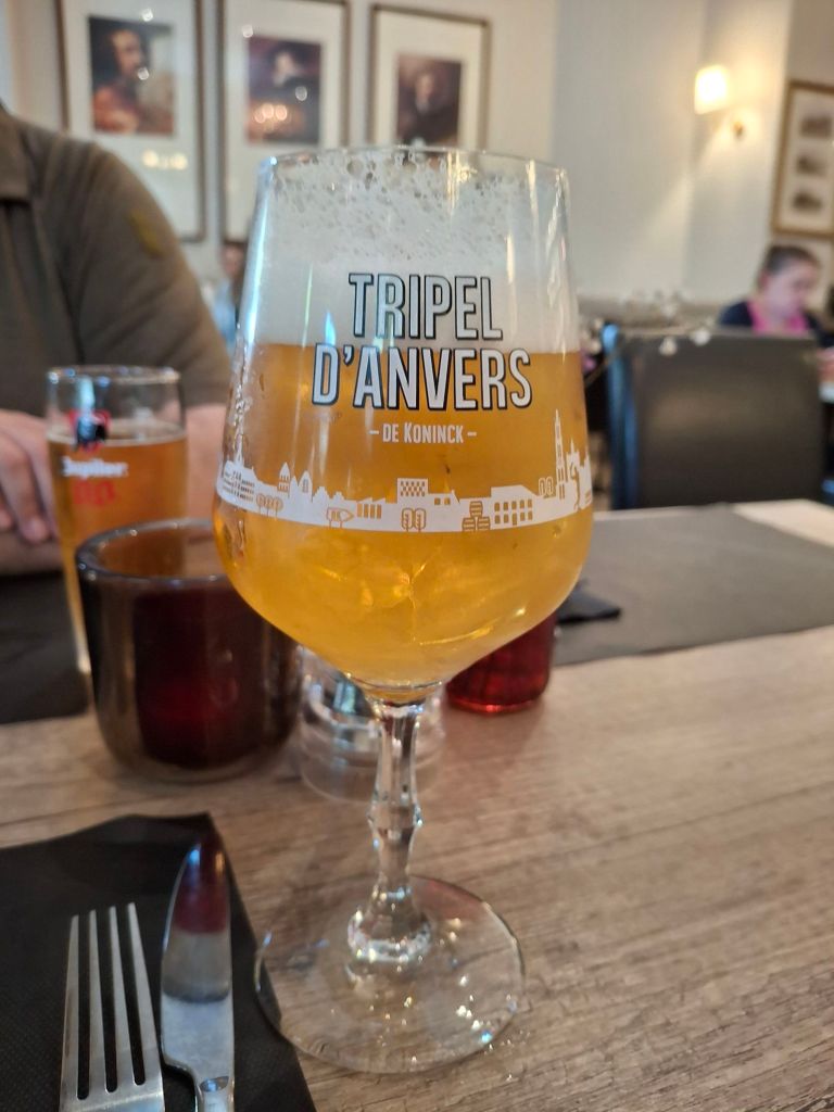 Large glass of light-coloured beer. The glass is patterned with a city skyline and has text "Tripel d'Anvers"