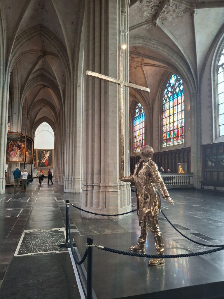 Inside a cathedral, a modern sculpture of a man in a suit carrying a cross by the base