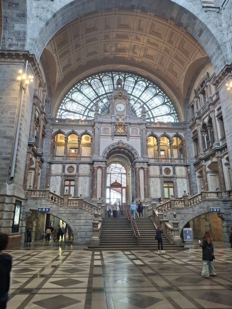 A railway station hall with a lot of grey stone columns, a huge semi-circular window at the top, and some gilt decorations. This is the same view as the picture in the jigsaw at the top of this post, but the frame is taller so we see more of the floor and ceiling. Also there are people in this shot.