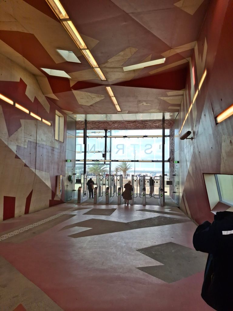 Automatic ticket barriers at a metro station; beyond them, sand and sea can be seen through the plate glass wall