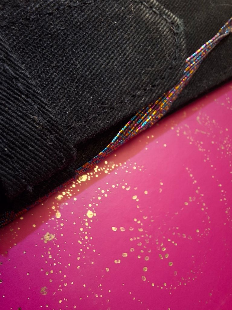 Close-up shot of bright pink card scattered with gold dots and stars, with the edge of a black pencil case and a brightly coloured length of elastic 